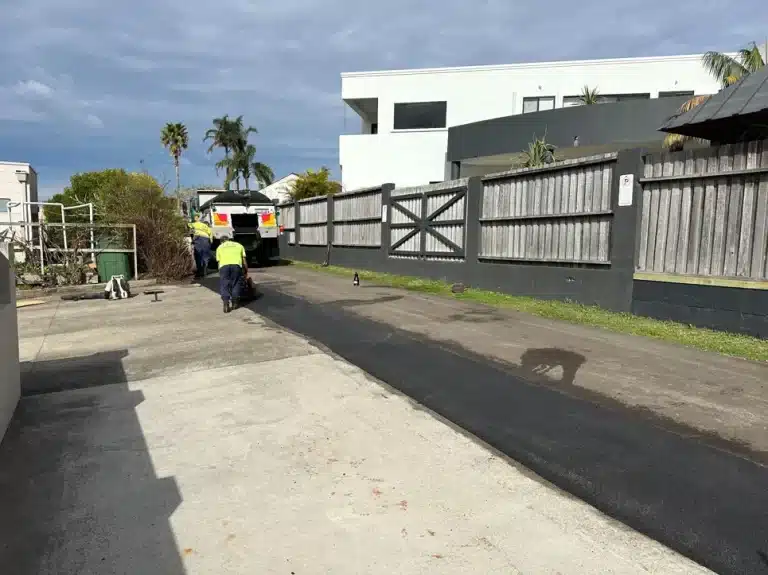Workers in high-visibility clothing apply fresh asphalt to a driveway between a building and a wooden fence, with industrial tools and a truck nearby.