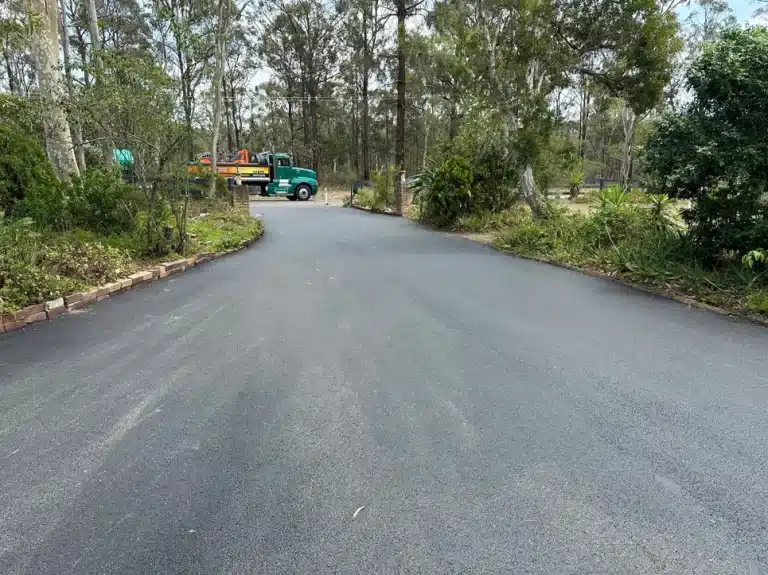 A newly paved asphalt road curves through a forested area with a green truck parked off the side.