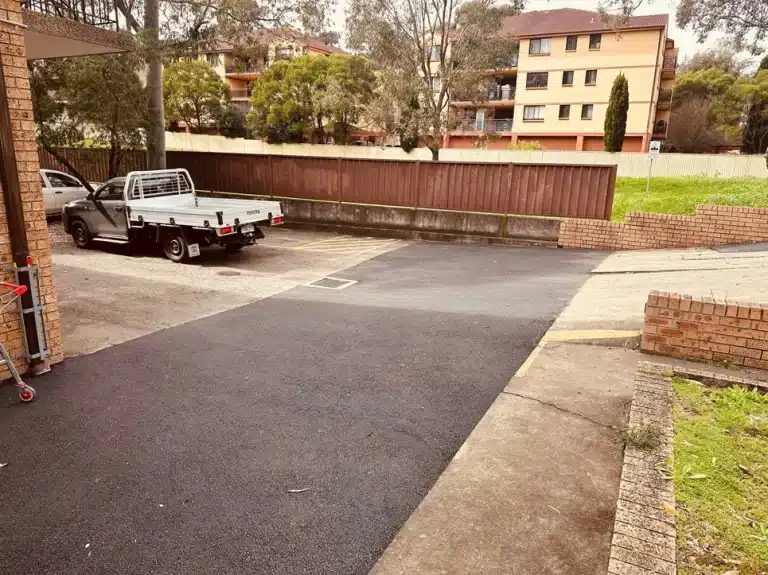 A parked flatbed truck sits in a small paved parking area near a wooden fence, with apartment buildings and trees in the background.