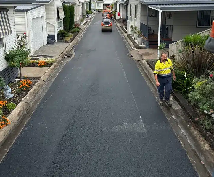 Workers in high-visibility clothing resurface a narrow residential street lined with mobile homes and gardens.